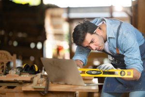 Un artisan menuisier utilise un niveau jaune pour calibrer un projet sur une table d'atelier couverte d'outils traditionnels et d'un ordinateur portable ouvert